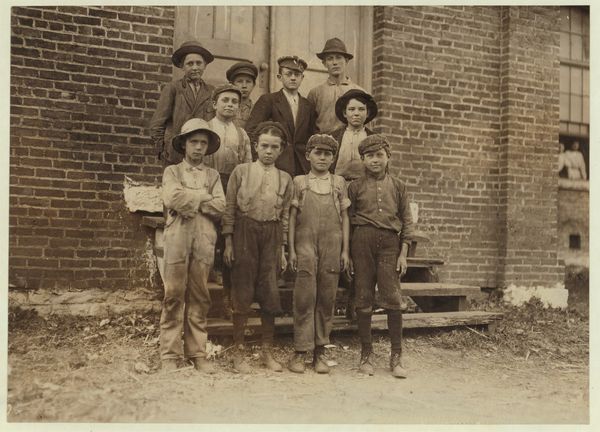Black-and-white photo of a group of young boys, child mill workers, standing against a brick wall