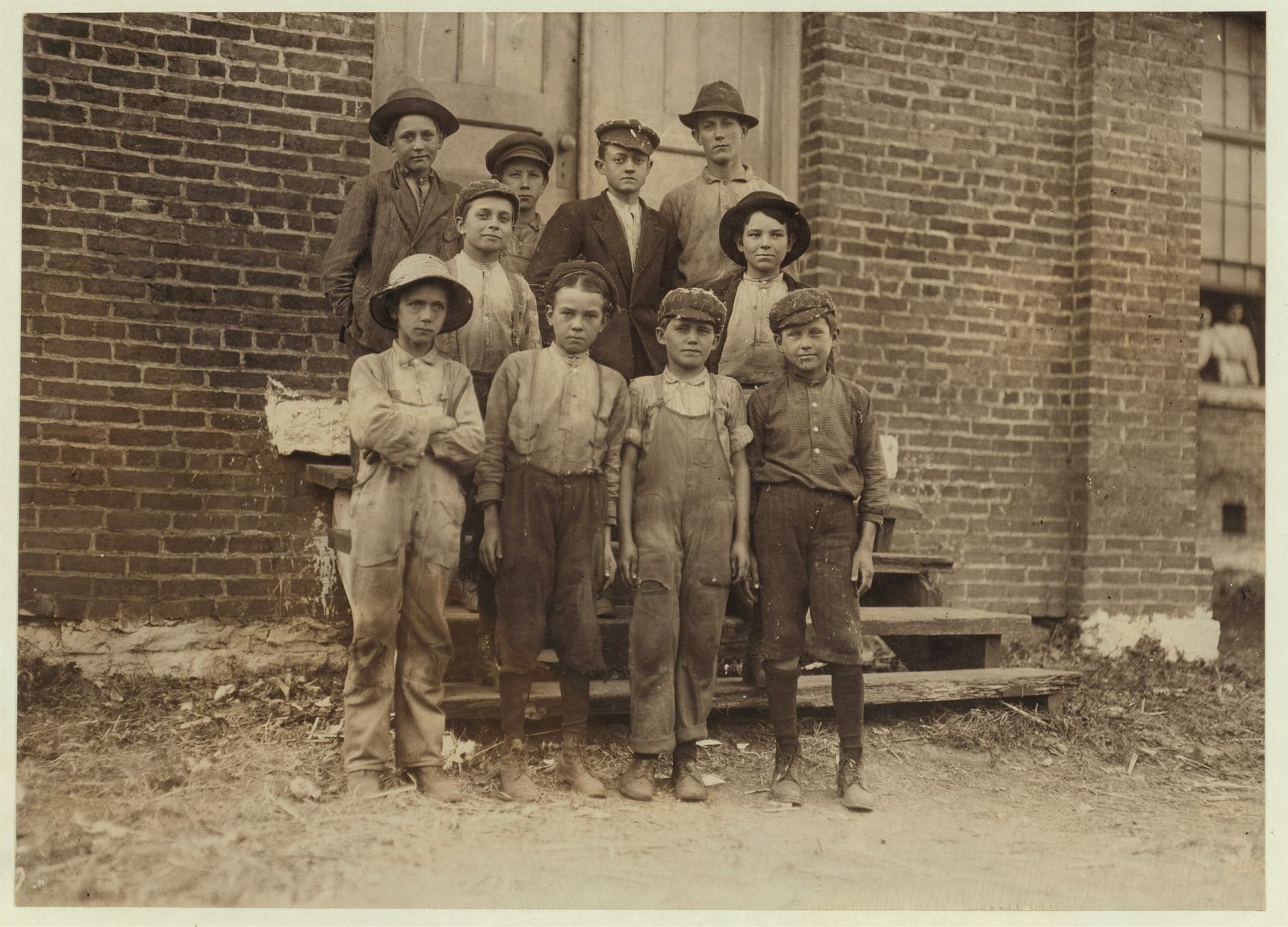 Black-and-white photo of a group of young boys, child mill workers, standing against a brick wall