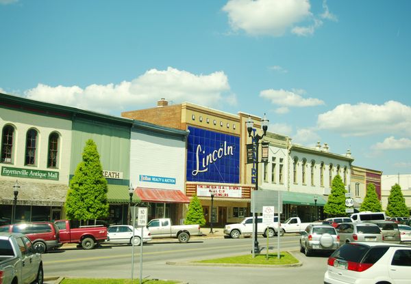 Two-story brick storefronts and the Lincoln Theater sign along the north side of the Fayetteville square on a sunny day