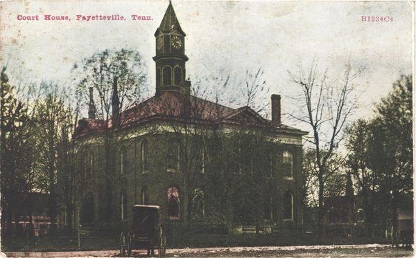 Tinted postcard of the 1874 Lincoln County Courthouse, a two-story brick building with a tall clock tower, behind bare trees with a horse carriage in front, about 1909