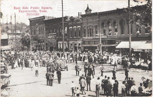 Old postcard of a crowd, a marching band and an early automobile in front of a row of two- and three-story brick stores on the east side of the Fayetteville square, about 1911