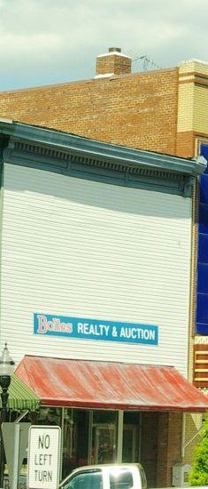 Two-story storefront with white metal siding over the upper floor, a red awning and a blue Bolles Realty & Auction sign, College St. E., 2015