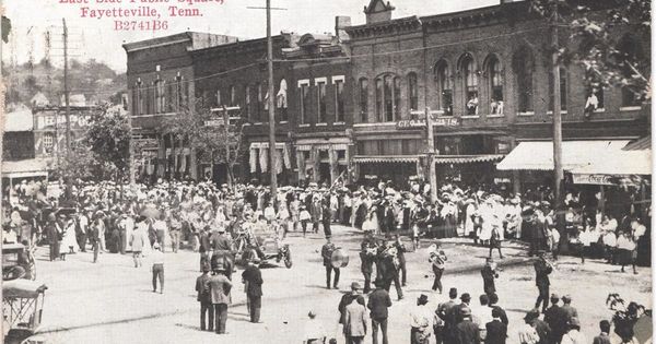 Old postcard of a crowd, a marching band and an early automobile in front of the brick stores on the east side of the Fayetteville square, about 1911